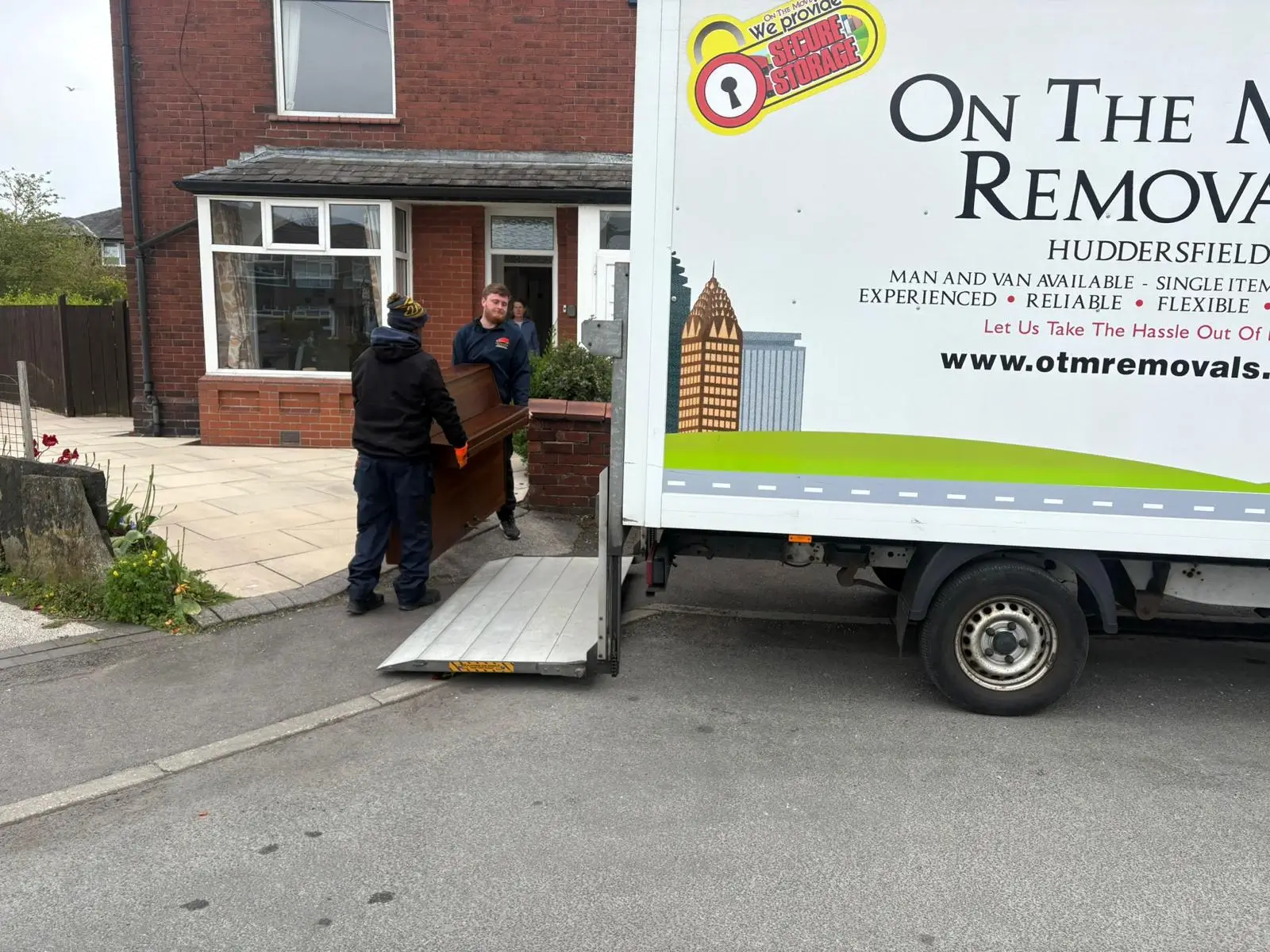 On The Move Removals two-person team walking a wrapped piano on a specialist dolly toward the van during a Huddersfield piano move